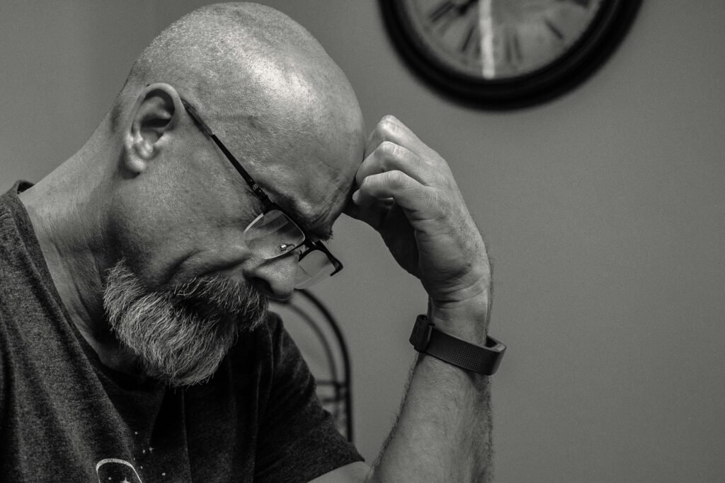 pexels-photo-1194196-1194196 Black and white portrait of a thoughtful bald man indoors, capturing a moment of reflection with a wall clock in the background.